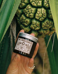 Hand holding a jar of 'Sol Medicinals' herbal salve in front of a pineapple plant.