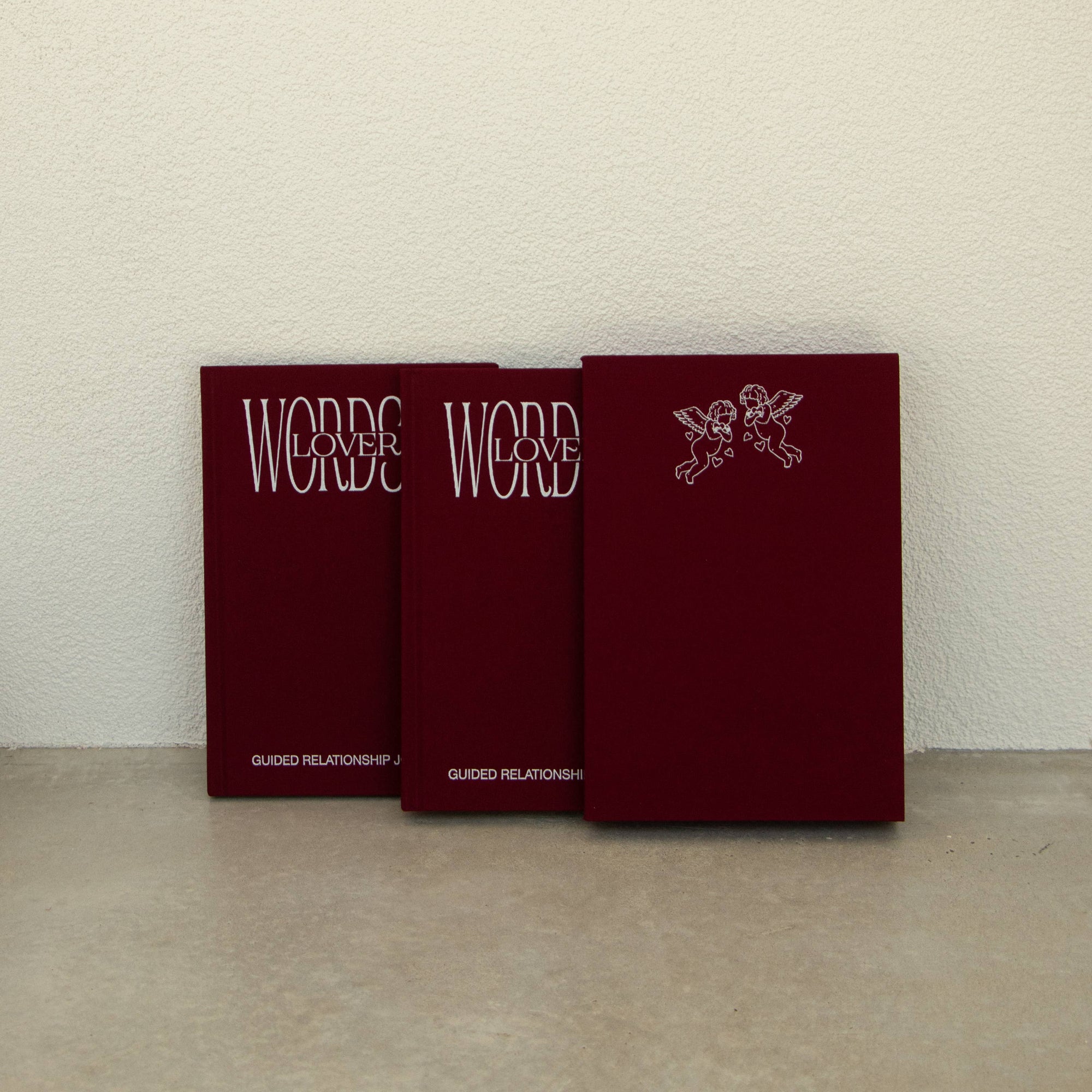 Three red books titled 'Words of Lovers' arranged in a row against a textured off-white wall and concrete floor.