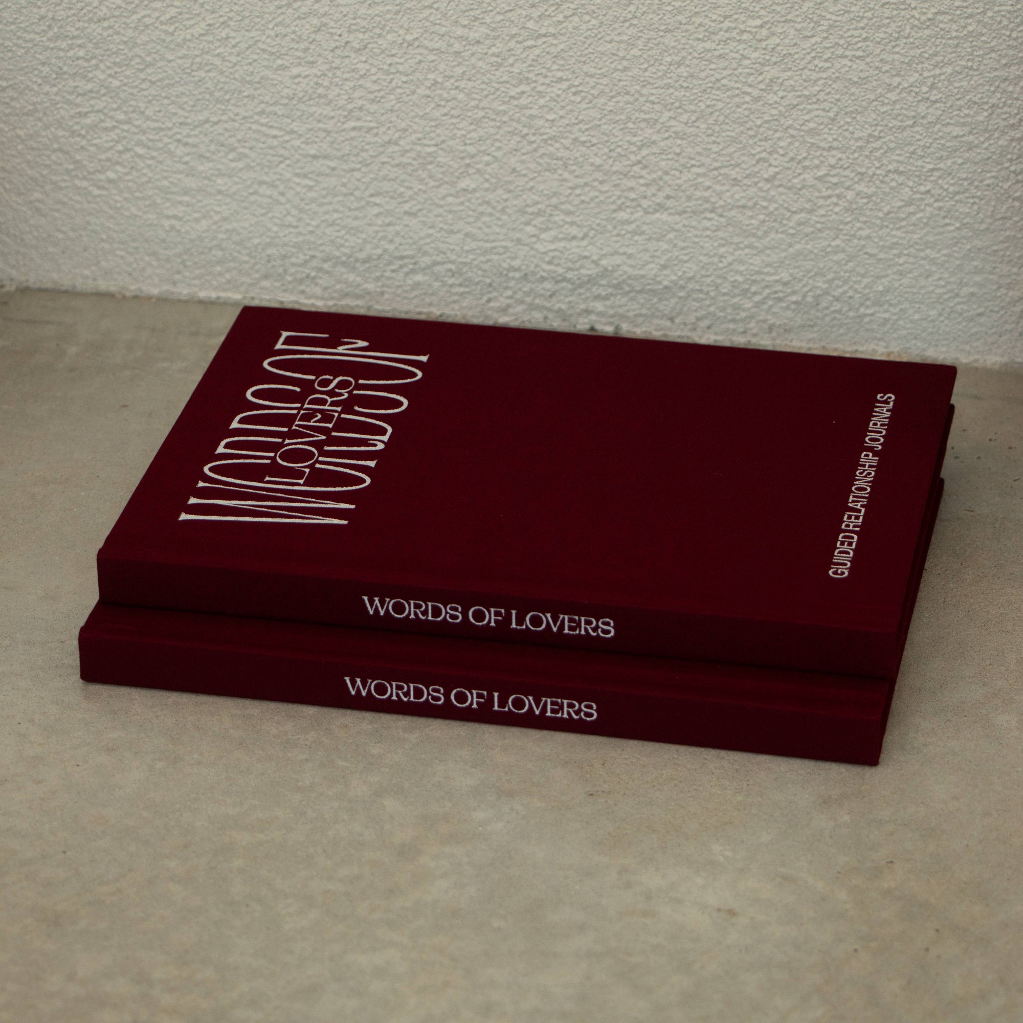 Two dark red hardcover books titled 'Words of Lovers' stacked on a light beige floor against a textured off-white wall.