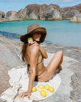 Woman in a bikini and wide-brimmed hat sitting on a rock with lemons, near turquoise water and rocky landscape.
